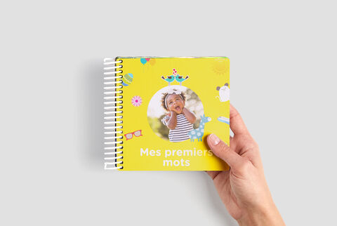 Une main tient un livre coloré avec le titre 'Mes premiers mots'. Le livre est jaune avec des illustrations amusantes.