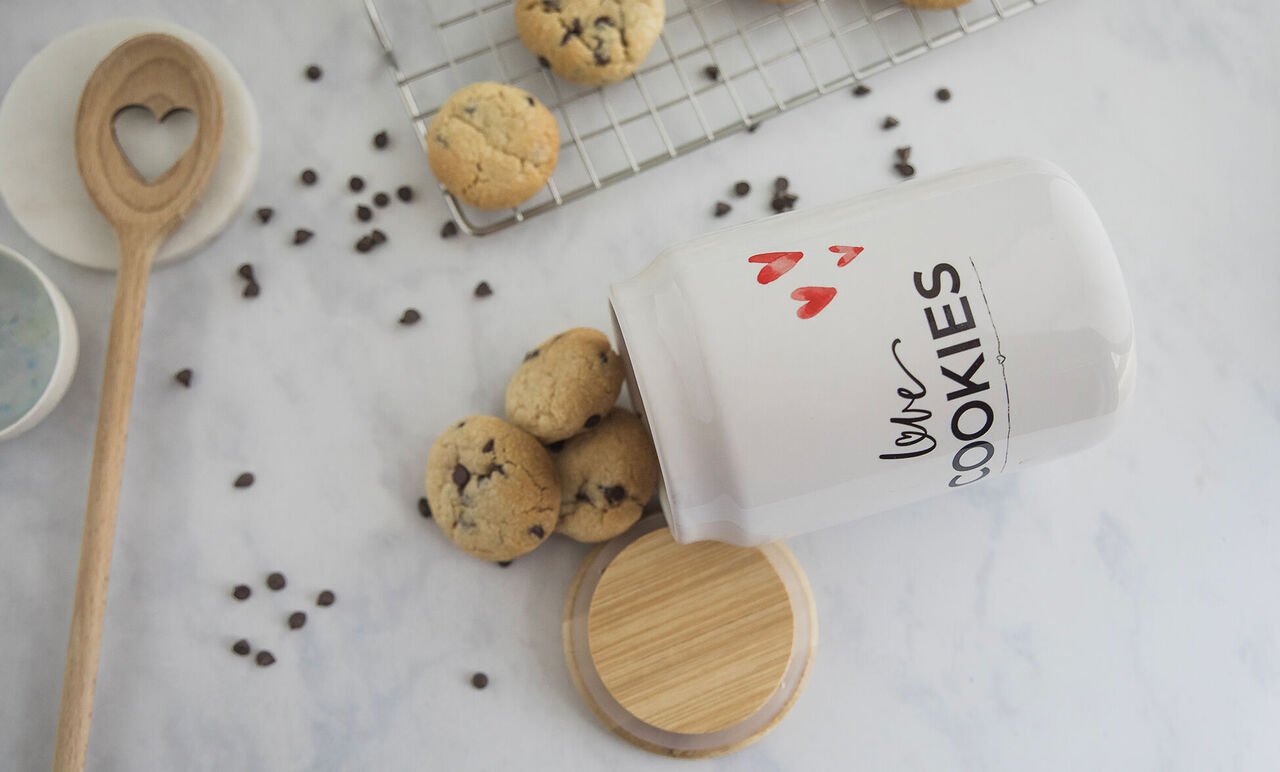 Pot à biscuits blanc renversé sur plan de travail clair avec inscription Love Cookies, couvercle en bois et cookies éparpillés.