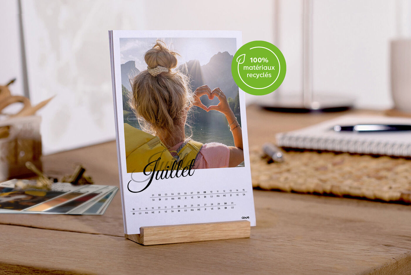 Une fille fait un geste de cœur devant une table. En arrière-plan, un calendrier. CEWE calendrier photo.
