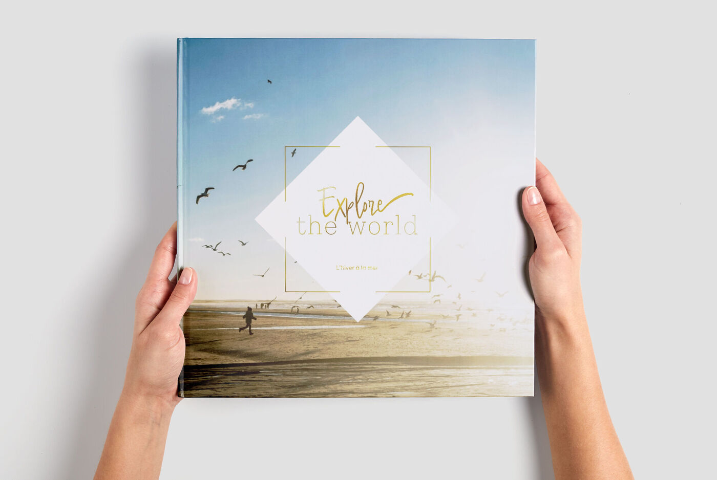 Quelqu'un tient un ALBUM PHOTO CEWE intitulé 'Explorer le Monde' sur une plage.