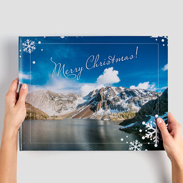 Quelqu'un tient un CEWE PhotoBook avec "Joyeux Noël" et un paysage hivernal en arrière-plan.