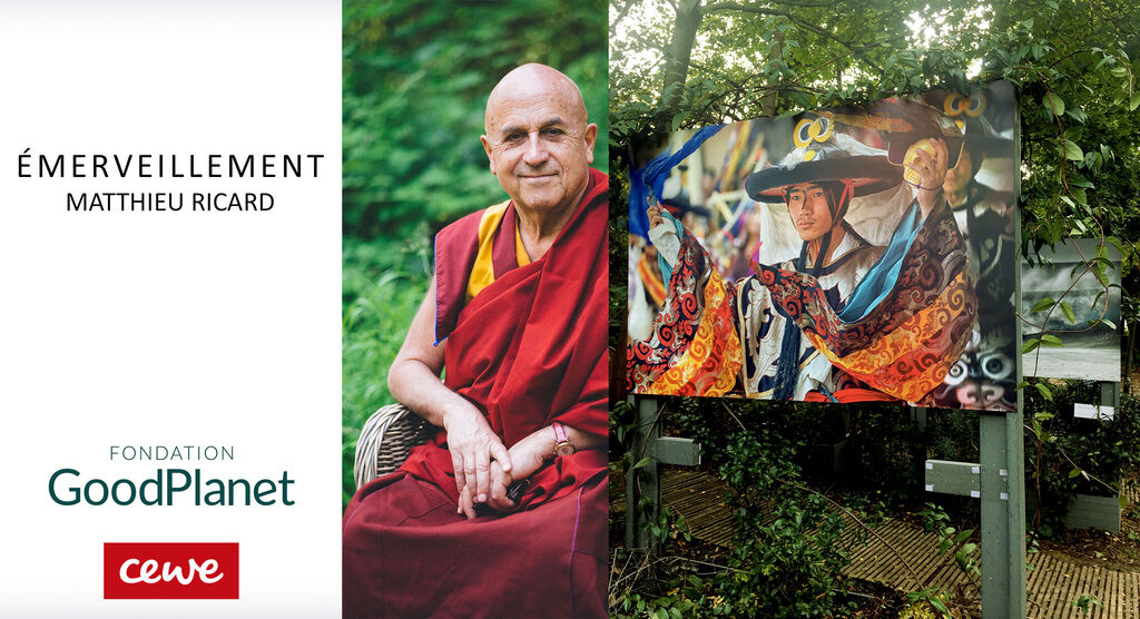 Un moine bouddhiste est assis à l'extérieur avec un sourire apaisant, regardant au loin. À droite, une affiche vibrante avec des motifs culturels.