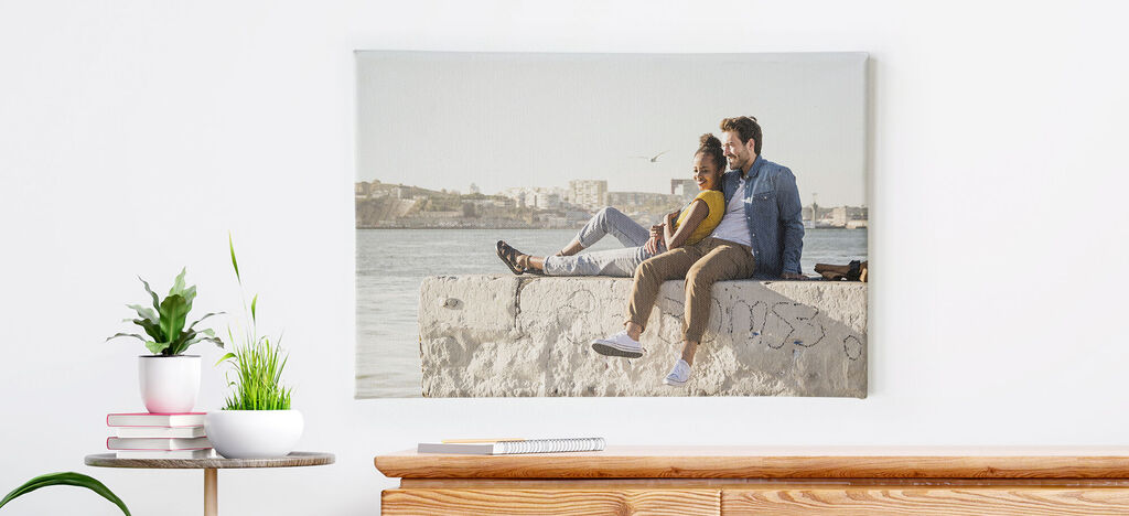 Un couple est assis confortablement au port sur un mur de pierre, sourit et admire la vue sur l'eau. En arrière-plan, des bâtiments sont visibles.