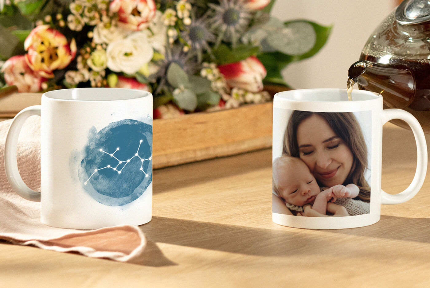 Deux tasses devant des fleurs en fleurs, l'une avec un motif floral, l'autre avec une photo de main de bébé.