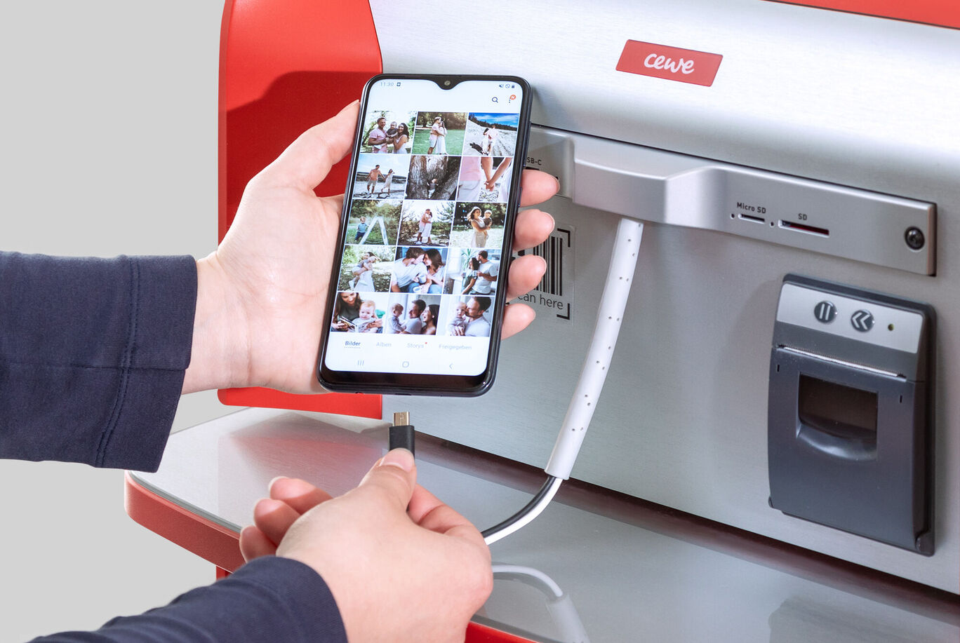 Une main connecte un smartphone à une station CEWE pour imprimer des photos dans un environnement lumineux