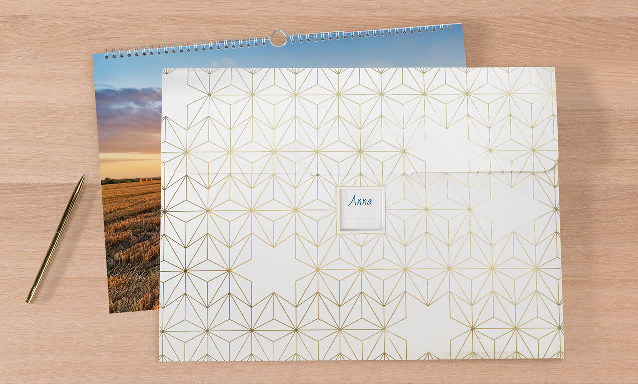 Un calendrier photo CEWE fermé sur une surface en bois, accompagné d'une photo et d'un stylo doré.
