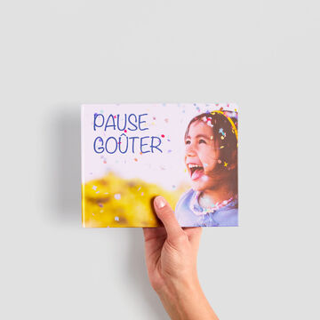 Une main tient un album photo CEWE avec le mot "Pause" et des confettis colorés en arrière-plan.