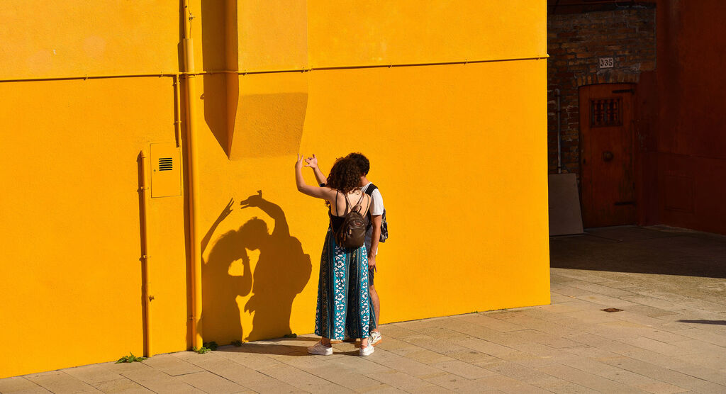 Un couple se tient devant un mur jaune soleil et voit l'ombre au soleil.
