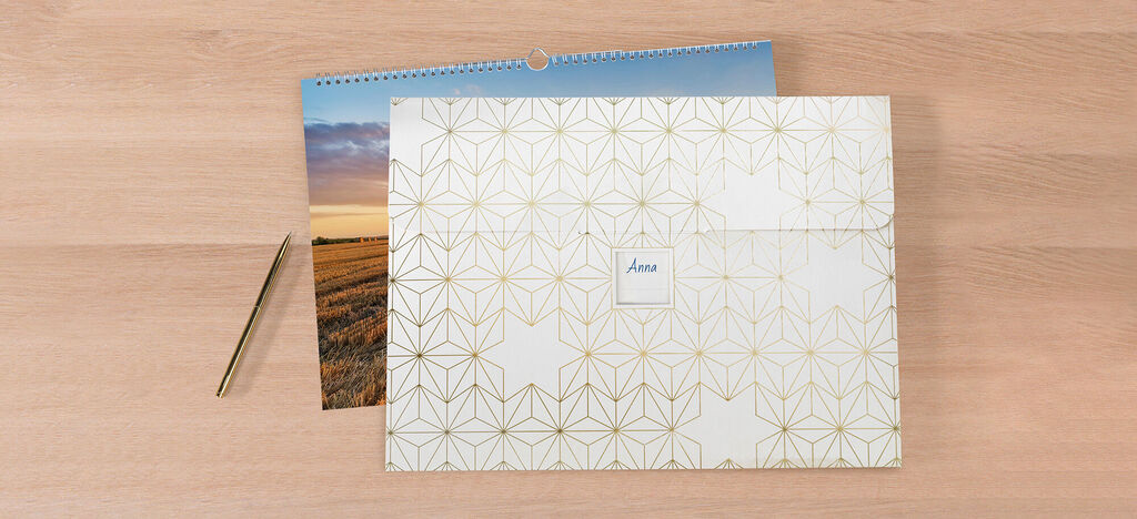 Des cartes de vœux CEWE élégantes avec un calendrier sur une table, stylos dorés à côté.