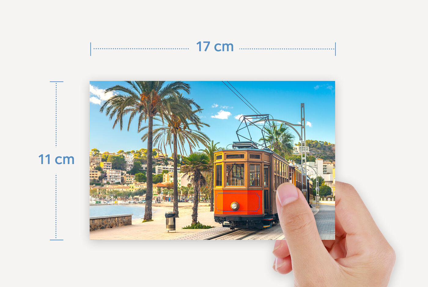 Une personne tient une carte postale avec un tram coloré dans une ville côtière. Des palmiers et la mer sont visibles en arrière-plan.